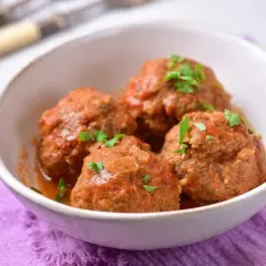 Pyszne pulpety w sosie pomidorowym bez smażenia, posypane natką pietruszki, podane w białej miseczce na fioletowej serwetce.