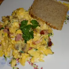 Pyszna jajecznica z boczkiem, pieczarkami i szczypiorkiem, udekorowana natką pietruszki, podana z kromką chleba.