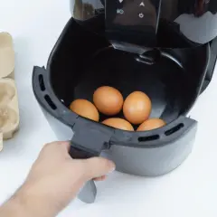 Ręka wkłada jajka do frytkownicy beztłuszczowej. Obok leży wytłaczanka. Szybkie i zdrowe jajka z airfryer.