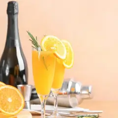 Dwa kieliszki orzeźwiającej mimosa z plasterkami pomarańczy i gałązką rozmarynu, obok butelki szampana i shakera.