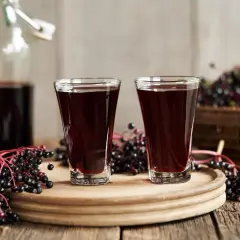 Dwa kieliszki pełne ciemnego, aromatycznego soku z czarnego bzu, obok gałązka z owocami i butelka.