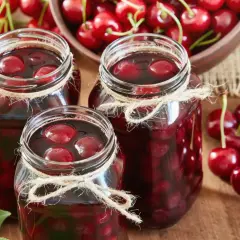 Słoiki z domowym kompotem z wiśni, ozdobione sznurkiem. Obok świeże wiśnie i miska pełna owoców.