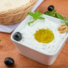 Jak zrobić tzatziki? Kremowy dip z jogurtu, czosnku i ziół, polany oliwą, z oliwkami i orzechami. Obok pieczywo.