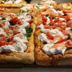 Dwie prostokątne pizze z burratą, pomidorkami i łososiem lub tuńczykiem. Puszyste ciasto, kremowy ser i świeże dodatki kuszą swoim wyglądem.