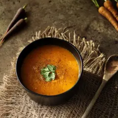 Pomarańczowa zupa z marchwi, idealna na lekki obiad. Obok miski leży drewniana łyżka i świeże marchewki.