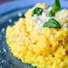 Aksamitny, żółty ryż do risotto, posypany parmezanem i udekorowany listkami bazylii, na niebieskim talerzu.