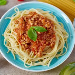 Pyszne spaghetti bolognese z passaty, podane na turkusowym talerzu, ozdobione listkiem bazylii. Obok czosnek, pomidorki i kolorowy pieprz.