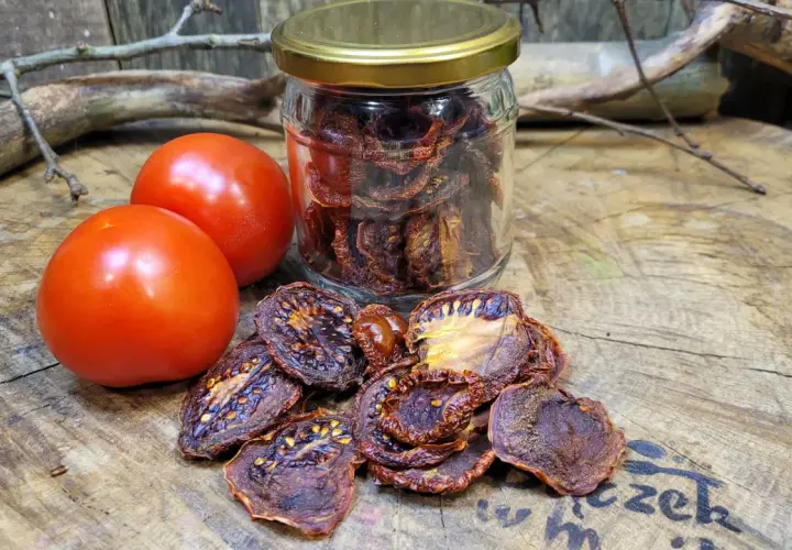 Słoik pełen aromatycznych suszonych pomidorów obok świeżych pomidorów i gałązek na drewnianym blacie.