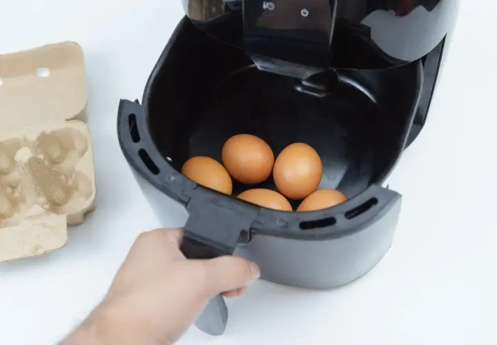 Ręka wkłada jajka do frytkownicy beztłuszczowej. Obok leży wytłaczanka. Szybkie i zdrowe jajka z airfryer.