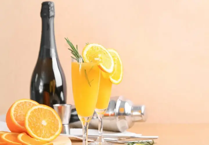 Dwa kieliszki orzeźwiającej mimosa z plasterkami pomarańczy i gałązką rozmarynu, obok butelki szampana i shakera.