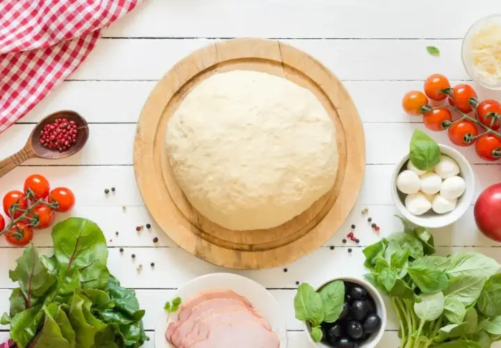 Świeże ciasto na pizzę czeka na składniki. Zastanawiasz się, ile może leżeć ciasto na pizze? Oto składniki na pyszną pizzę: pomidorki, mozzarella, oliwki, bazylia, szynka.
