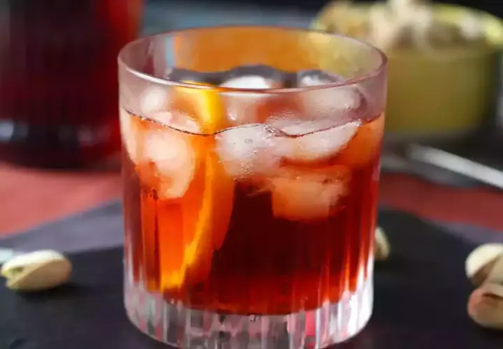 Klasyczny negroni przepis: czerwony koktajl z lodem i pomarańczą w szklance, obok pistacje.