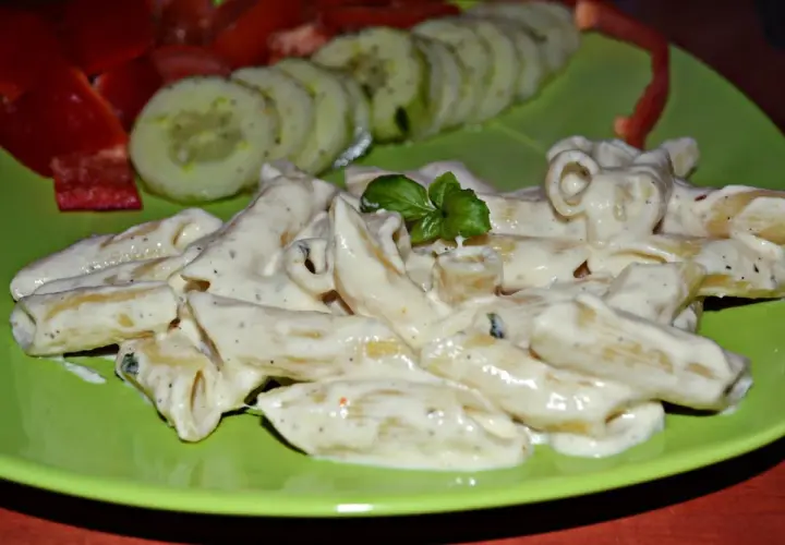 Penne w kremowym sosie z mozzarellą, udekorowane bazylią. Obok plasterki ogórka i papryka.