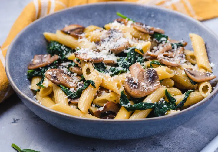 Penne z pieczarkami i szpinakiem, posypane parmezanem, w szarej misce.