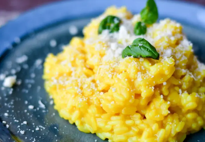 Aksamitny, żółty ryż do risotto, posypany parmezanem i udekorowany listkami bazylii, na niebieskim talerzu.