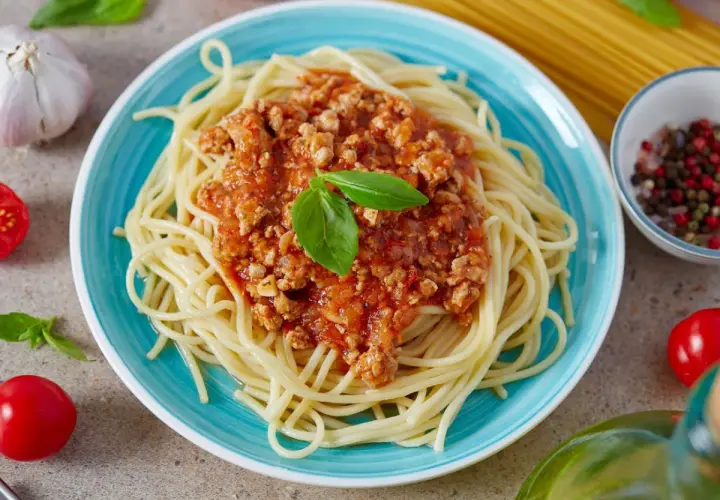 Pyszne spaghetti bolognese z passaty, podane na turkusowym talerzu, ozdobione listkiem bazylii. Obok czosnek, pomidorki i kolorowy pieprz.