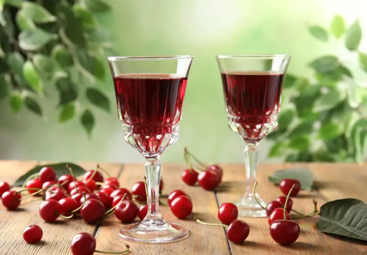 Dwa kieliszki z rubinową, szybką nalewką z wiśni na spirytusie, otoczone świeżymi wiśniami na drewnianym stole.