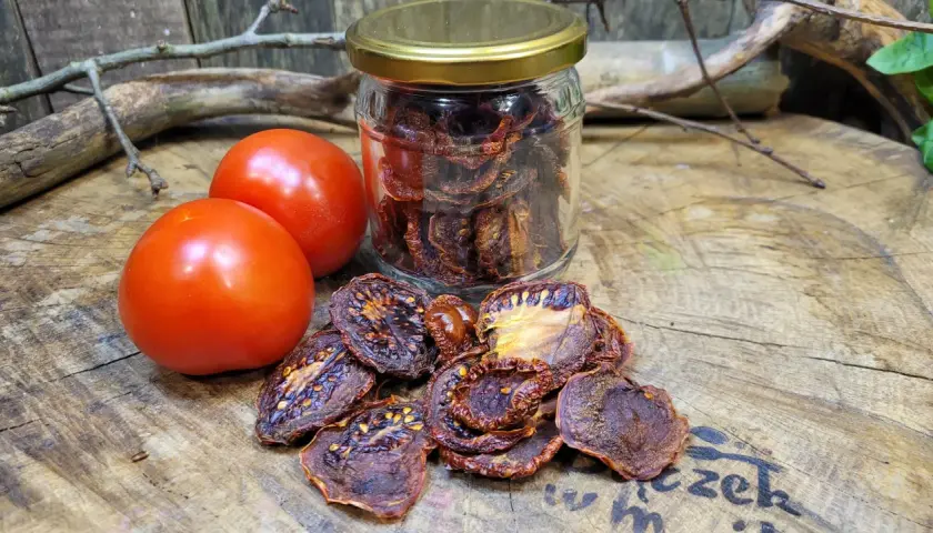 Słoik pełen aromatycznych suszonych pomidorów obok świeżych pomidorów i gałązek na drewnianym blacie.