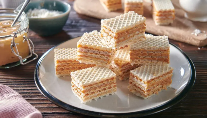 Pyszne wafle z kajmakiem z puszki, ułożone na talerzu. Obok słoiczek z kremowym kajmakiem i miseczka z wiórkami kokosowymi.