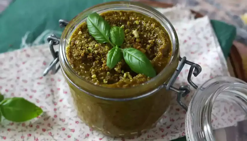 Słoik z domowym pesto pistacjowym, ozdobiony listkami bazylii i posypany pistacjami.