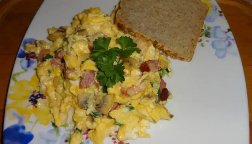 Pyszna jajecznica z boczkiem, pieczarkami i szczypiorkiem, udekorowana natką pietruszki, podana z kromką chleba.