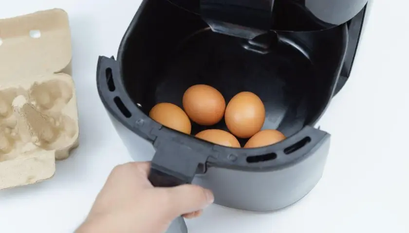 Ręka wkłada jajka do frytkownicy beztłuszczowej. Obok leży wytłaczanka. Szybkie i zdrowe jajka z airfryer.