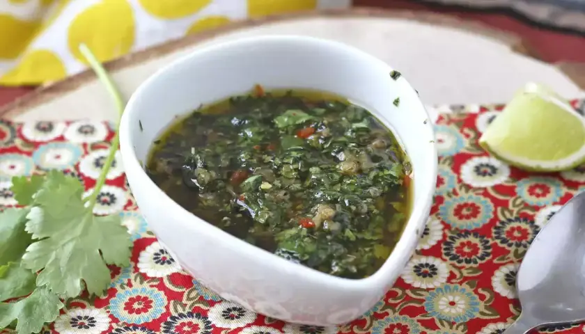 Aromaticzny sos chimichurri w białej miseczce, obok ćwiartka limonki i świeża kolendra.