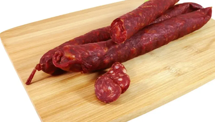 Trzy długie kiełbaski chorizo leżą na drewnianej desce. Jedna jest przekrojona, ukazując charakterystyczny, czerwony środek.