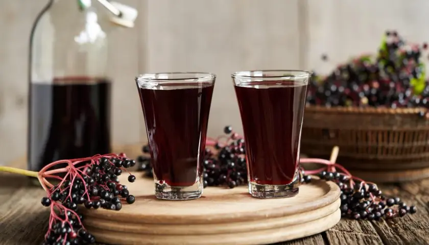 Dwa kieliszki pełne ciemnego, aromatycznego soku z czarnego bzu, obok gałązka z owocami i butelka.