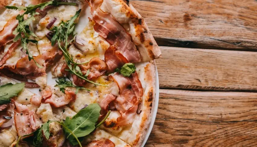 Pyszna pizza carbonara z chrupiącym boczkiem i rukolą, podana na drewnianym blacie.