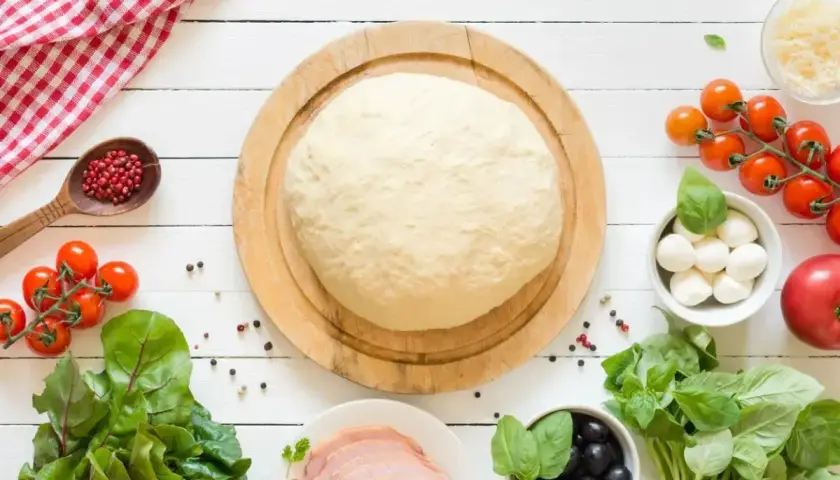 Świeże ciasto na pizzę czeka na składniki. Zastanawiasz się, ile może leżeć ciasto na pizze? Oto składniki na pyszną pizzę: pomidorki, mozzarella, oliwki, bazylia, szynka.