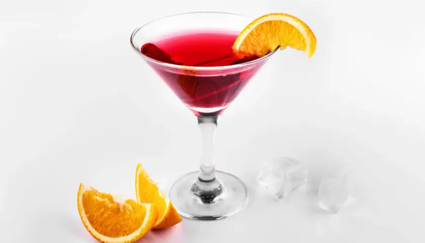 Elegancki, czerwony drink cosmopolitan z plasterkiem pomarańczy, obok kostki lodu i pomarańczy.