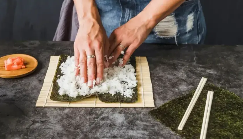 Ręce rozprowadzają ryż na nori, przygotowując zaprawę do sushi. Obok leżą pałeczki i marynowany imbir.