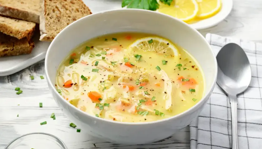 Aromatyczna zupa cytrynowa z kurczakiem, marchewką i makaronem, ozdobiona plasterkiem cytryny i natką pietruszki. Podana z chlebem.