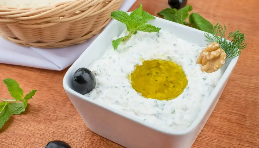 Jak zrobić tzatziki? Kremowy dip z jogurtu, czosnku i ziół, polany oliwą, z oliwkami i orzechami. Obok pieczywo.