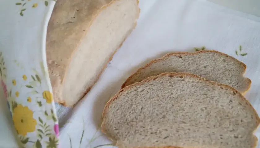 Dwa rodzaje chleba: bochenek owinięty w kwiecisty materiał i dwa kromki na białym obrusie.