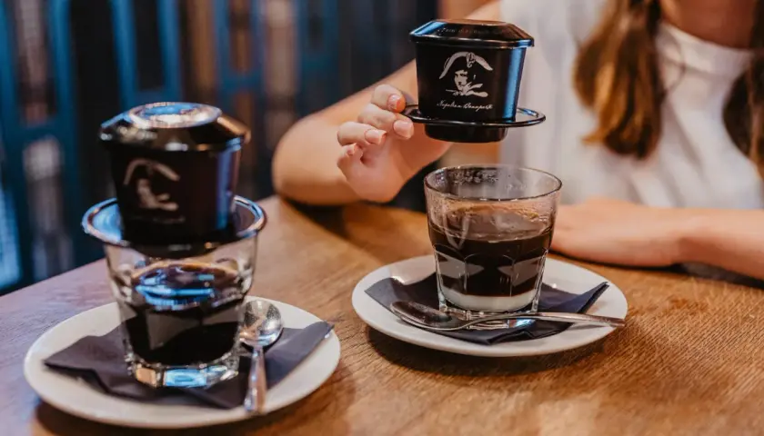 Przygotowanie kawy po wietnamsku. Kobieta trzyma zaparzacz, a obok stoją dwa szklanki z kawą i mlekiem skondensowanym.