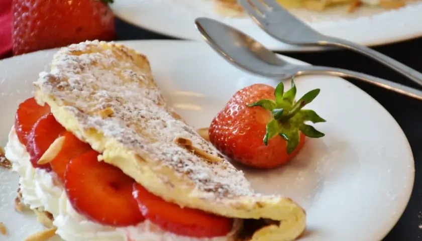 Puszysty omlet z bitą śmietaną i truskawkami, posypany cukrem pudrem i płatkami migdałów. Obok leży świeża truskawka.