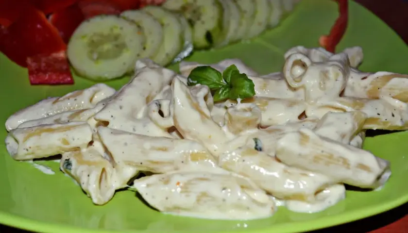 Penne w kremowym sosie z mozzarellą, udekorowane bazylią. Obok plasterki ogórka i papryka.