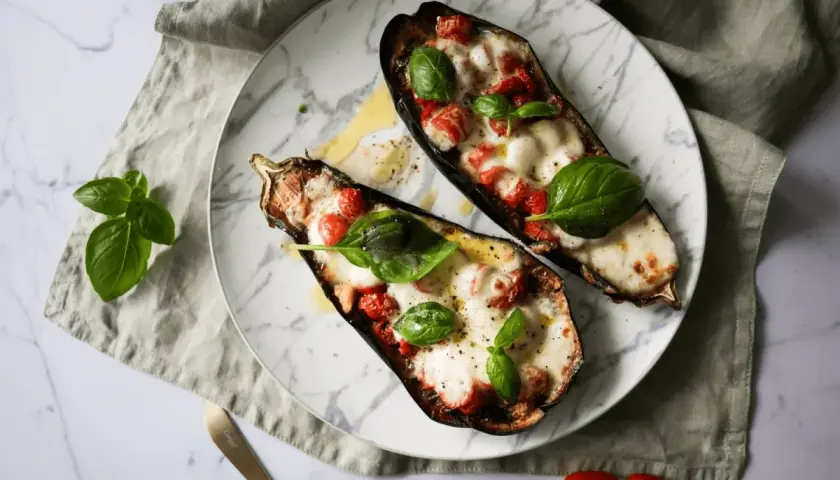 Dwa połówki bakłażana pieczonego z pomidorkami i mozzarellą, ozdobione listkami bazylii.
