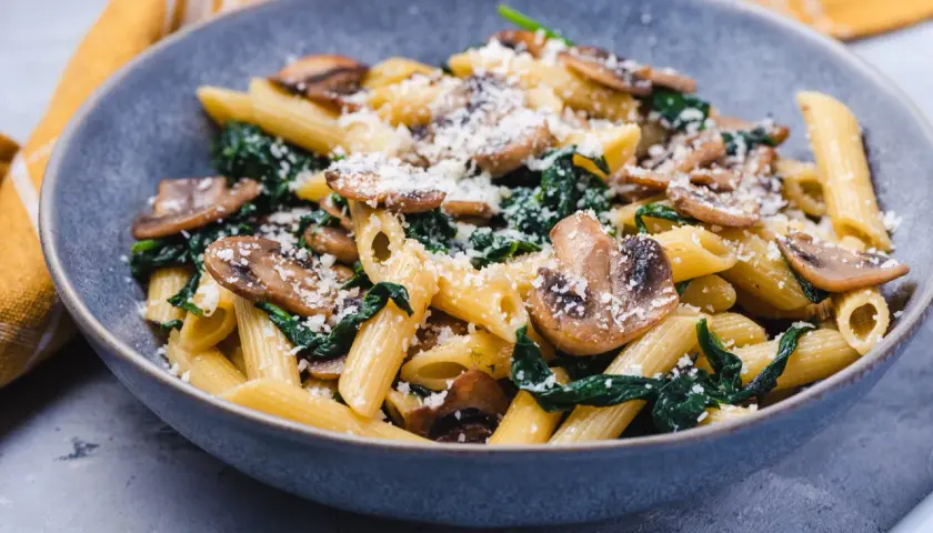 Penne z pieczarkami i szpinakiem, posypane parmezanem, w szarej misce.