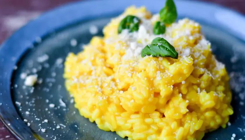 Aksamitny, żółty ryż do risotto, posypany parmezanem i udekorowany listkami bazylii, na niebieskim talerzu.