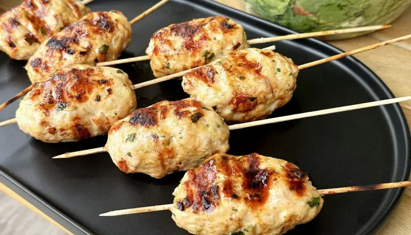 Szaszłyki z mięsa mielonego, apetycznie przypieczone na grillu, leżą na czarnym talerzu. Obok widać miskę z sałatką.