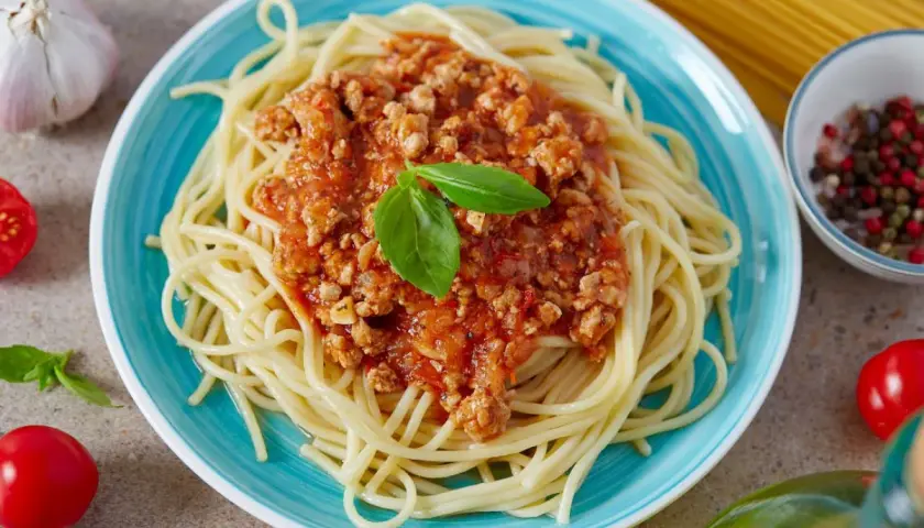 Pyszne spaghetti bolognese z passaty, podane na turkusowym talerzu, ozdobione listkiem bazylii. Obok czosnek, pomidorki i kolorowy pieprz.