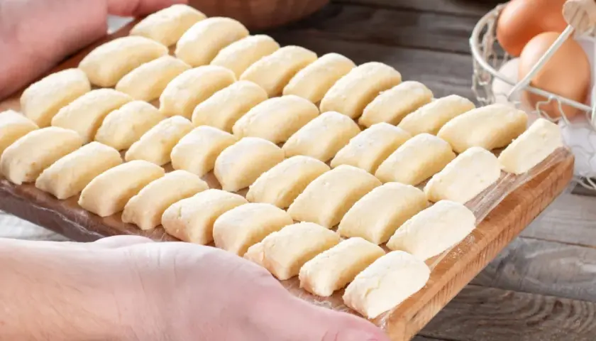 Gotowe do gotowania kluski leniwe, przepis babci. Na desce ułożone rzędy pierożków z twarogu, obok jajka i twaróg.