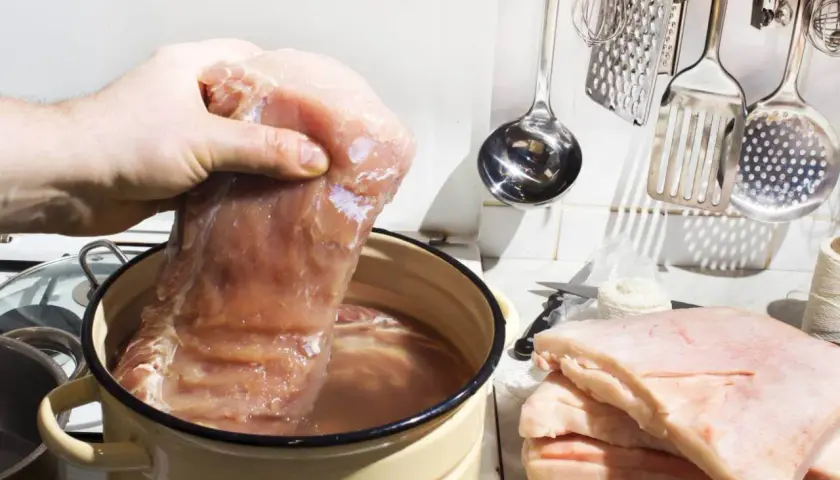 Przygotowanie solanki do mięsa: ile soli na litr wody? Duży kawałek mięsa zanurzany w wodzie w garnku, obok stos słoniny i kuchenne akcesoria.