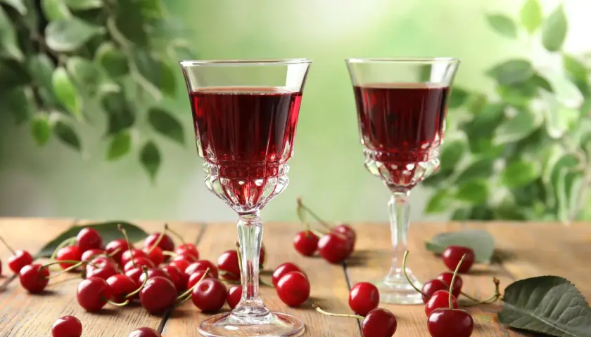 Dwa kieliszki z rubinową, szybką nalewką z wiśni na spirytusie, otoczone świeżymi wiśniami na drewnianym stole.