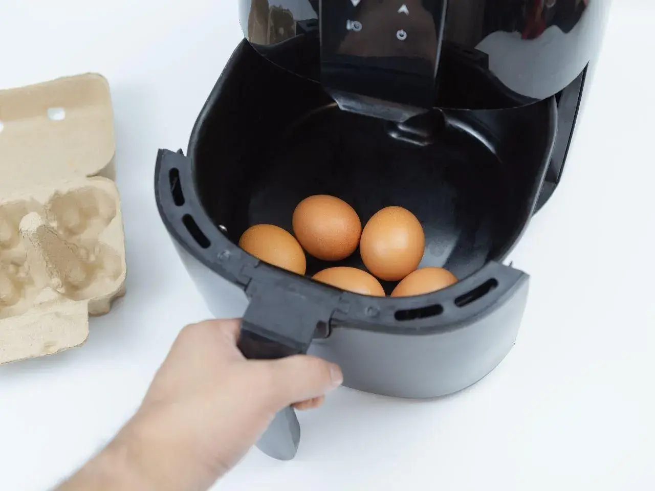 Ręka wkłada jajka do frytkownicy beztłuszczowej. Obok leży wytłaczanka. Szybkie i zdrowe jajka z airfryer.