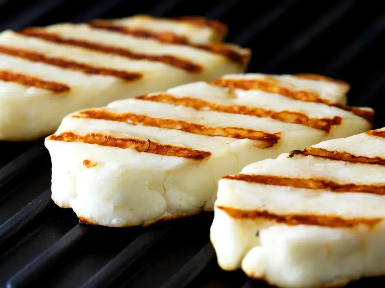 Trzy kawałki halloumi z grilla, z apetycznymi paskami, leżą na czarnej, grillowej powierzchni.
