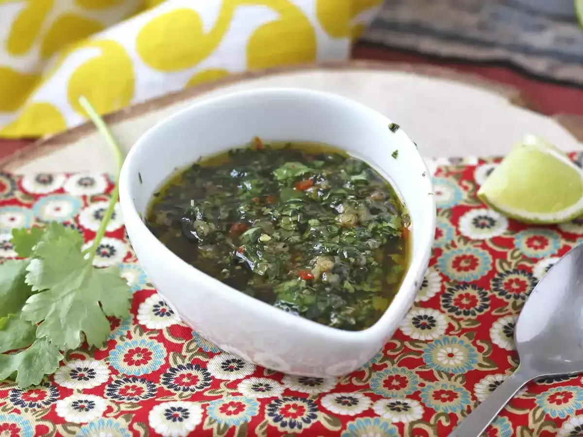 Aromaticzny sos chimichurri w białej miseczce, obok ćwiartka limonki i świeża kolendra.
